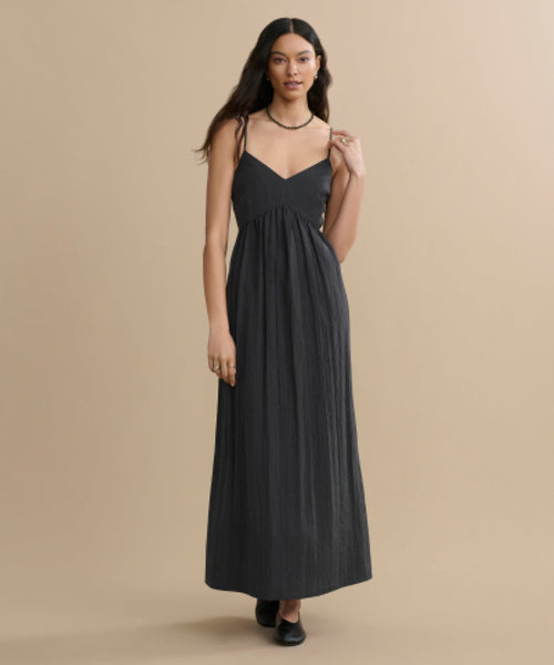 A woman stands against a beige background wearing the Alyssandra Dress, a long black party dress with thin straps and a V-neckline. She has long, wavy hair, and pairs her outfit with a black necklace and black shoes.