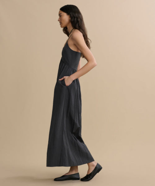 A woman with long dark hair stands in profile against a beige background, wearing the Alyssandra Dress—a sleeveless, ankle-length dark gray dress with pockets, made from textured fabric, paired with black slip-on shoes.