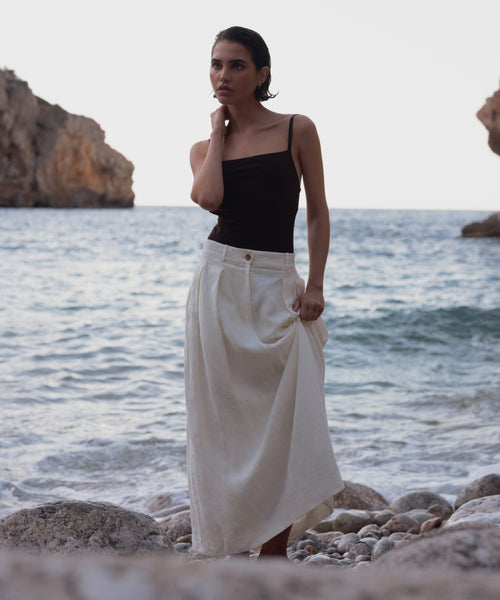 Wearing the Annabel Skirt and a black sleeveless top, a woman stands on a rocky beach with waves and cliffs behind her. She holds one side of the skirt, gazing thoughtfully into the distance.