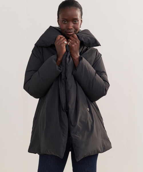 A person wearing the Apres Coat—a puffy, oversized black down coat with a high collar—stands against a plain light background, holding the collar close to their face.