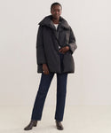 A woman models the Apres Coat—black down outerwear—paired with dark blue jeans, brown ankle boots, and a white top against a light background, her hand in her pocket for an effortlessly stylish winter look.