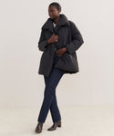 Wearing the Apres Coat, dark blue jeans, and brown ankle boots, a woman stands against a light background, gazing to the side with one hand on her jacket—showcasing effortless winter essentials style.