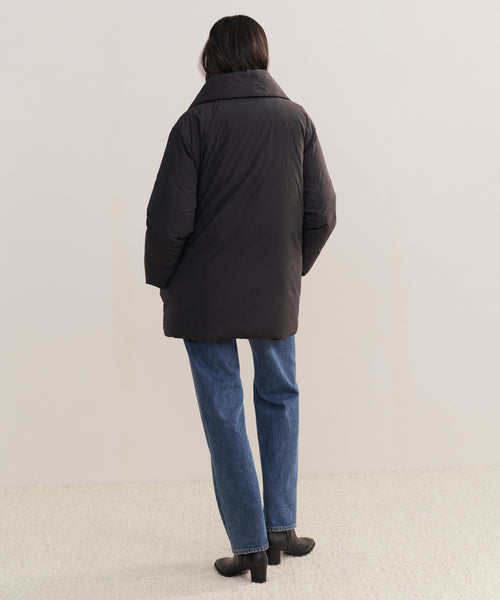 A person with long dark hair, facing away, wears the Apres Coat—this season’s must-have black down jacket—paired with blue jeans and black boots on a light surface against a plain, light background.