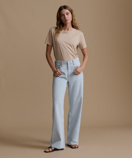 Woman wearing a beige t-shirt and light blue jeans against a brown background
