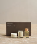 The Aromatherapy Bath Set features a brown box behind three items: hydrating body balm, a glass candle, and an oil bottle with a wooden cap, all displayed on a neutral surface.