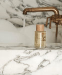 A bottle of bath oil from the Aromatherapy Bath Set rests on a white marble tubs edge, as water pours from a brass faucet and bubbles fill the background.