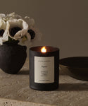 The Aspen Glass Candle, crafted from a natural wax blend, rests on a stone surface beside a black vase with white flowers and a dark bowl, adding cozy elegance to the scene.