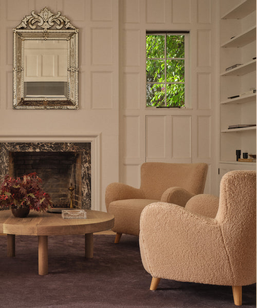 A cozy living room with two Aspen Chairs in oatmeal sherpa, a round wood coffee table topped with red leaves, a decorative fireplace, ornate mirror, built-in shelves, and a window framing lush greenery outside.