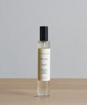 Aspen Room Spray, a clear bottle from jenni kayne with seasonal scents like birchwood, rests on a wooden surface against a plain gray background.