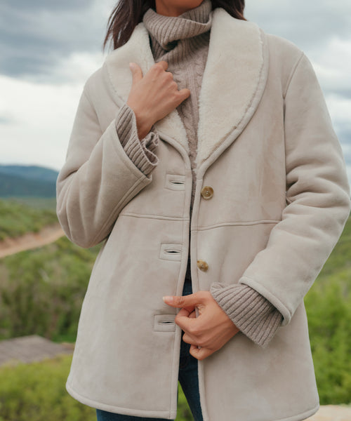 A woman stands outdoors against a blurred green landscape, effortlessly showcasing the Aspen Shearling Coat in light beige with button details over a taupe turtleneck. Her face is not visible.