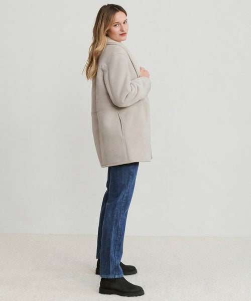 A woman with long hair stands sideways against a plain light background, looking over her shoulder while wearing the Aspen Shearling Coat, blue jeans, and black shoes.