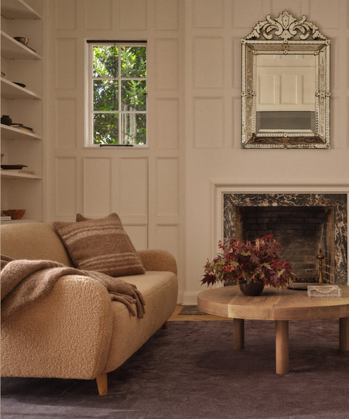 A cozy living room features the Aspen Sofa in oatmeal sherpa, a brown throw and pillows, a round wood coffee table with a vase of dark red leaves, a fireplace with a mirror above it, and a window framing green outdoor scenery.