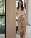 A woman with long brown hair, wearing the Avery Crewneck striped sweater and loose tan pants, stands indoors by a wooden wall, smiling at the camera. A window behind her reveals greenery outside.