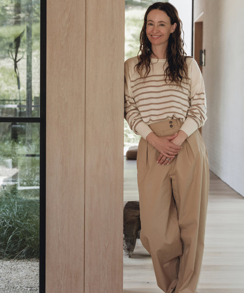A woman with long brown hair, wearing the Avery Crewneck striped sweater and loose tan pants, stands indoors by a wooden wall, smiling at the camera. A window behind her reveals greenery outside.