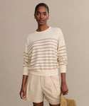 A woman stands against a beige background wearing the Avery Crewneck, a striped cotton-merino sweater with tan stripes, paired with light beige pleated shorts. She holds a textured tan clutch and wears small hoop earrings.
