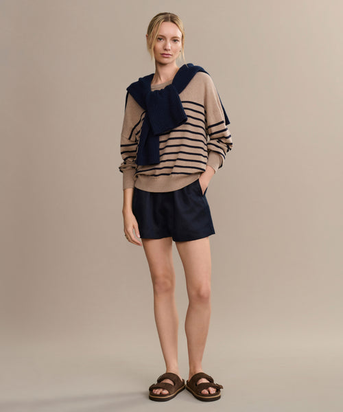 A woman stands against a neutral background wearing the Avery Crewneck striped sweater, with a navy sweater over her shoulders, navy shorts, and brown sandals. Hands in pockets, she exudes a relaxed coastal style.