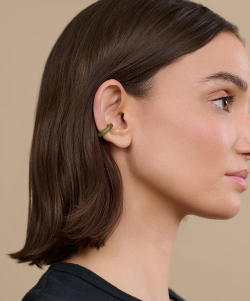 A woman with straight brown hair wears a small, green Bagel Ear Cuff on the outer edge of her left ear as she faces right against a beige background.