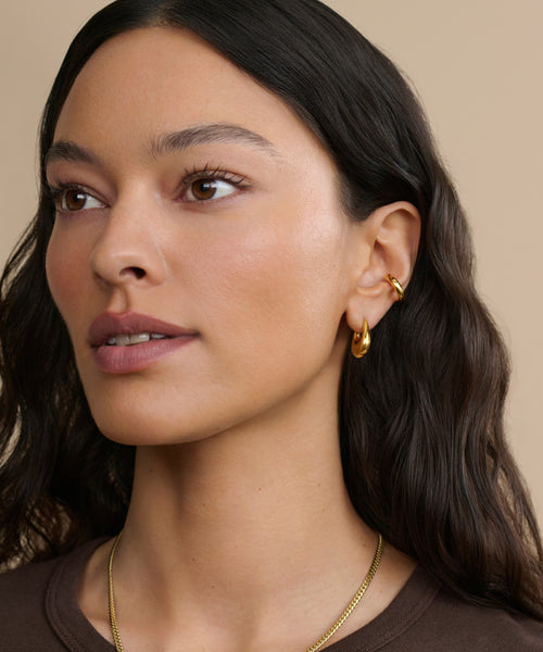A woman with long, wavy dark hair wears a Bagel Ear Cuff and gold hoop earrings, paired with a gold necklace. She poses in natural makeup and a brown top against a neutral beige background, showcasing an everyday modern jewelry look.