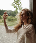 A woman with curly hair and closed eyes sprays her face with Balancing Mist while standing by an open door, wearing a light textured blouse. Green trees and a gravel path are visible outside.