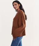 A woman with long dark hair models the Barrett Cardigan with blue jeans, standing sideways and looking at the camera against a plain light background.