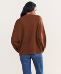 A person with long dark hair is seen from behind wearing the oversized Barrett Cardigan in brown knit with blue jeans, standing against a plain light background.