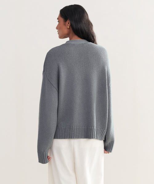 A person with long dark hair wears the loose-fitting, gray Barrett Cardigan and white pants, standing with their back to the camera against a light, neutral background.