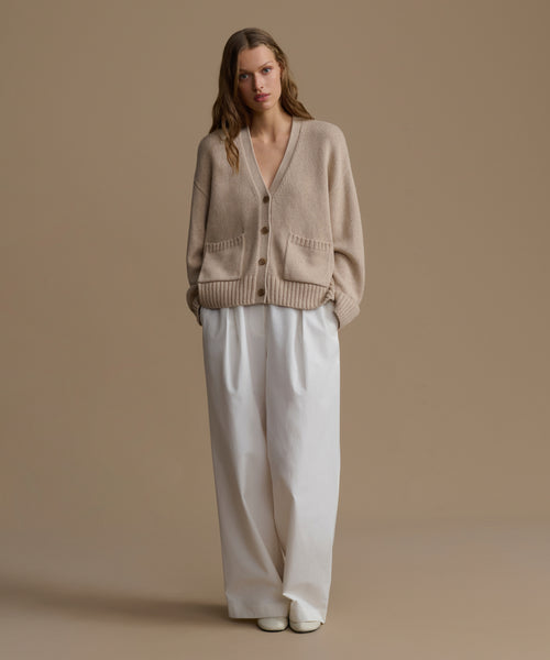 Woman wearing a beige cashmere cardigan and white pants on a brown background