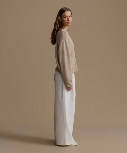 Woman wearing a beige cashmere sweater and white pants against a brown background