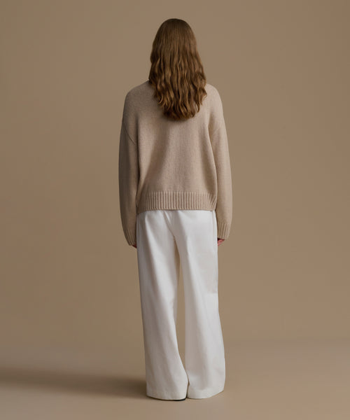 Person wearing a beige cashmere sweater and white pants against a brown background