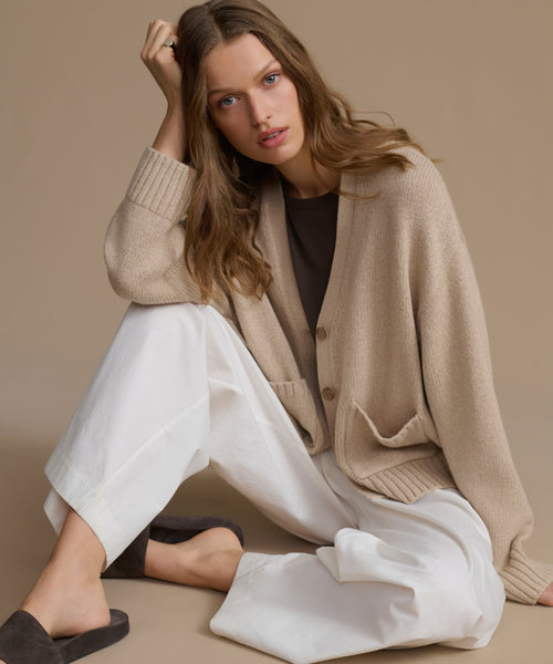 Woman wearing a beige cashmere cardigan and white pants sitting on a beige surface.