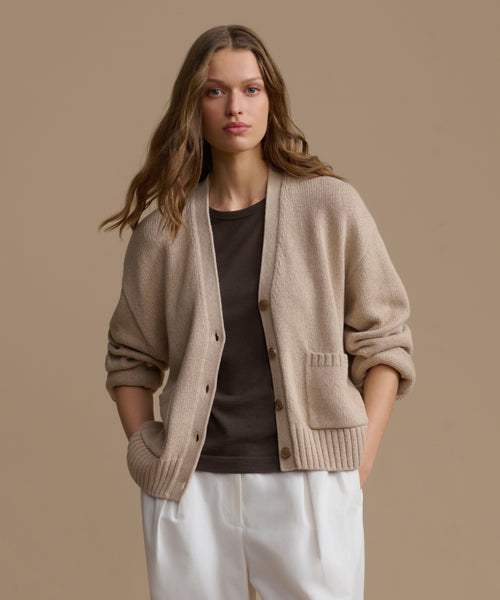 Woman wearing a beige cardigan over a dark top and white pants on a beige background
