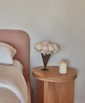 A wooden bedside table holds a Basil Glass Candle, a metal vase with pale pink flowers, and stands beside a bed with a light pink upholstered headboard and crisp white bedding.