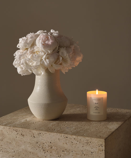 A white ceramic vase with white and pale pink flowers sits on a stone surface next to a Basil Glass Candle in a glass jar, set against a plain beige background.