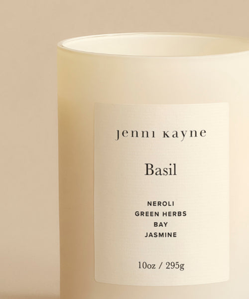 The Basil Glass Candle is a 10oz beige candle in a frosted jar labeled jenni kayne, hand-poured in Los Angeles with scent notes of neroli, green herbs, bay, and jasmine. Featured on Oprah’s Favorite Things.