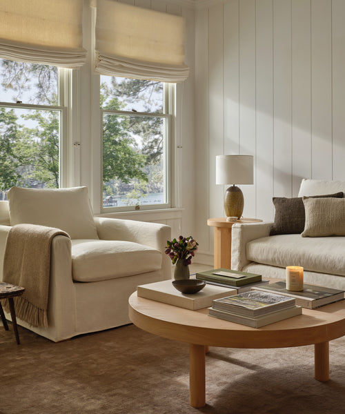 A cozy living room with cream armchairs, a Bay Coffee Table decorated with books and a candle, a side table with a lamp, and large windows with Roman shades that let in natural light and offer views of trees outside.