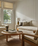 A cozy living room features cream sofas and a handcrafted Bay Coffee Table adorned with books, a candle, and flowers. Sunlight filters through windows with light Roman shades, creating a warm and inviting atmosphere.