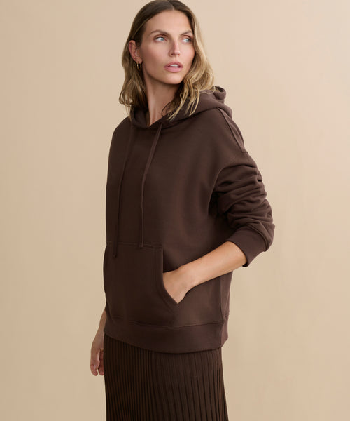 A person wearing the Bay Hoodie with a front pocket and a brown pleated skirt stands against a beige background, looking off to the side with one hand in the hoodie pocket.