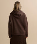 A person with light brown hair is seen from behind, wearing the Bay Hoodie in chocolate brushed fleece and a pleated brown skirt, standing against a plain beige background.