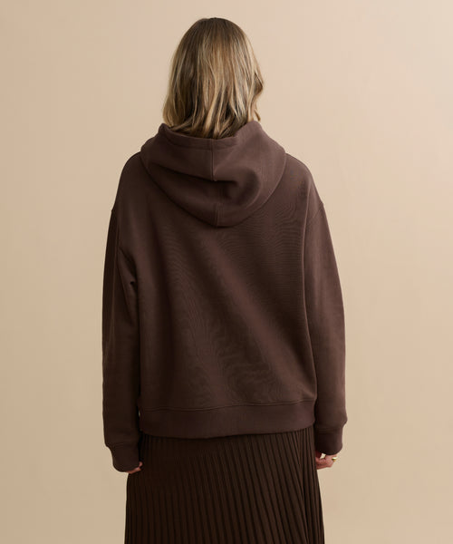 A person with light brown hair is seen from behind, wearing the Bay Hoodie in chocolate brushed fleece and a pleated brown skirt, standing against a plain beige background.