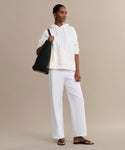 A woman stands against a beige background wearing the Bay Hoodie in ivory, white wide-leg pants, and black sandals. She carries a large black woven tote and wears small earrings—the perfect cozy wardrobe look.
