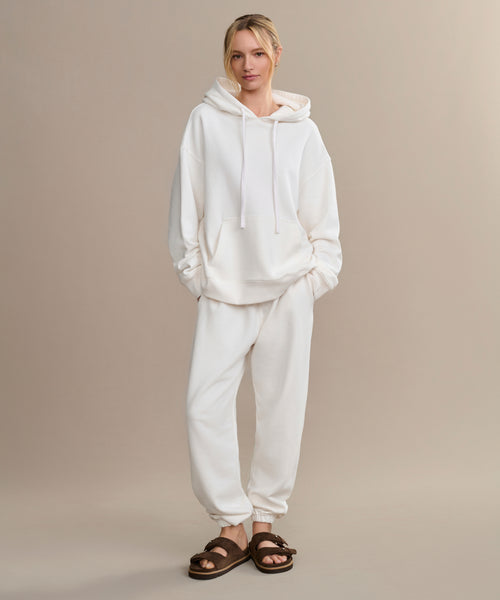A person stands against a beige background wearing an oversized Bay Hoodie in ivory, matching white sweatpants, and brown fuzzy sandals—perfect for a cozy wardrobe.
