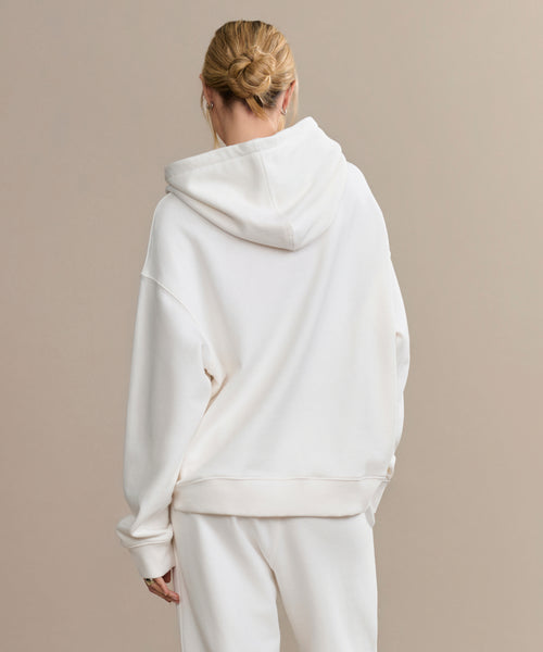 A person with blonde hair in a low bun, wearing the oversized Bay Hoodie in ivory and matching white pants, stands with their back to the camera against a plain beige backdrop—a cozy wardrobe essential.