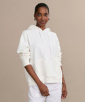 A woman stands against a beige background wearing the Bay Hoodie in ivory and white pants, looking at the camera with a slight smile and her hands relaxed by her sides—a perfect cozy wardrobe addition.