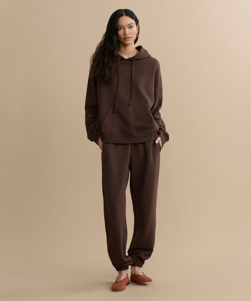A woman with long dark hair stands against a beige background wearing a matching brown brushed fleece hoodie, Bay Sweatpant, and brown slip-on shoes, with her hands in her pockets.