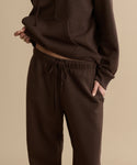 Model wears a matching dark brown brushed fleece Bay Sweatpant set, with one hand in their pocket and the other lifting the hoodie to reveal part of their midriff, set against a plain beige background.