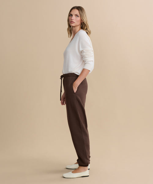 A woman with loose hair and a neutral expression stands in profile against a beige background, wearing a white long-sleeve top, Bay Sweatpant in brown, and white flat shoes.