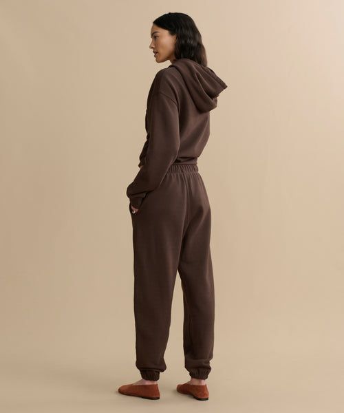 A person stands sideways, facing left, wearing a matching dark brown brushed fleece hoodie and Bay Sweatpant with hands in pockets, plus brown slippers, against a beige background.