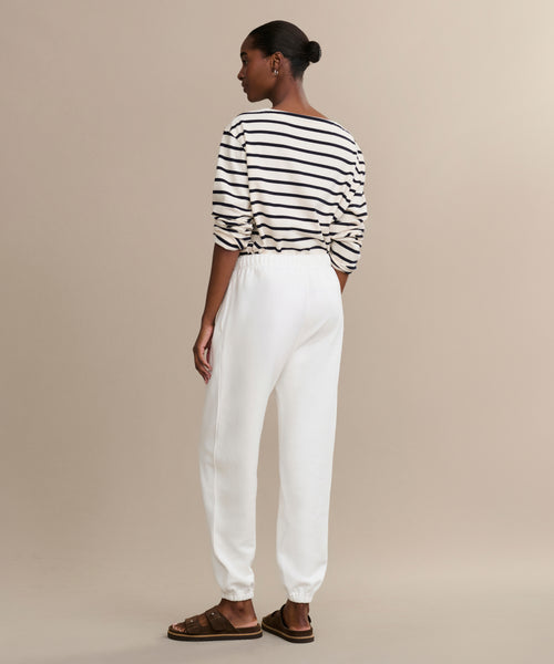 A person faces away, wearing a black-and-white striped long-sleeve shirt, Bay Sweatpant athletic sweats in white, and brown sandals against a plain beige background.