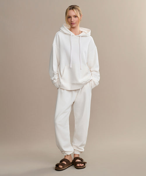 A person stands against a plain beige background, wearing an oversized white hoodie, the Bay Sweatpant in matching white brushed fleece, and brown fuzzy sandals.