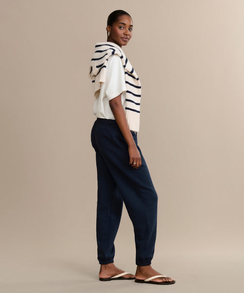 A woman stands sideways wearing a white shirt, navy Bay Sweatpant joggers, sandals, and a cream sweater with navy stripes draped over her shoulders. The background is plain beige.